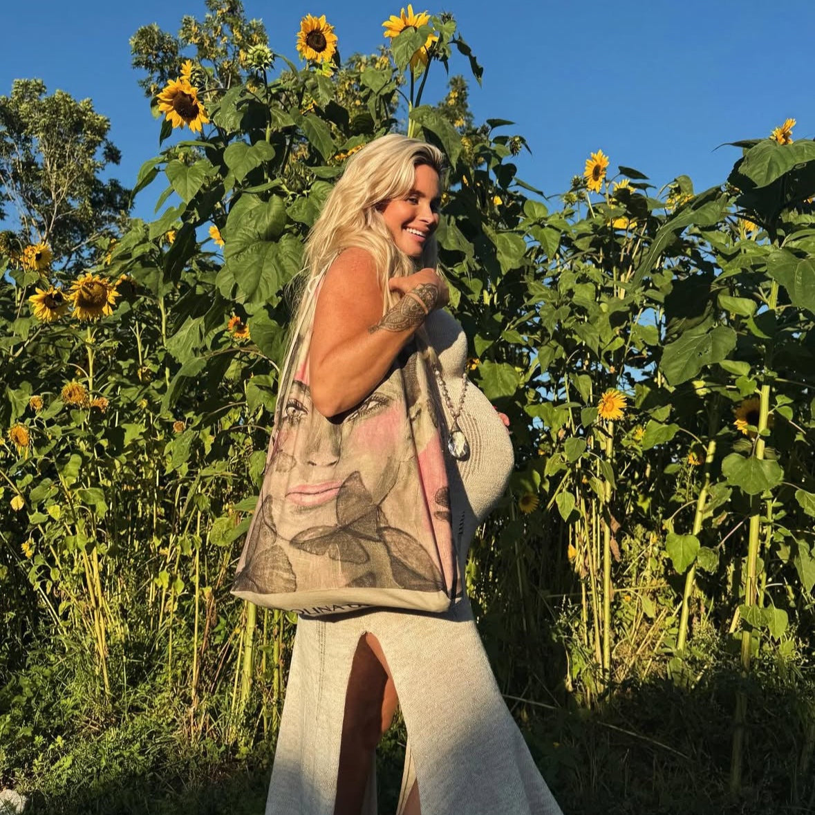 Carolina Gynning bär Boho tygväska Butterfly Queen i ljusrosa och grått över axeln i ett solrosfält, klädd i sommarklänning